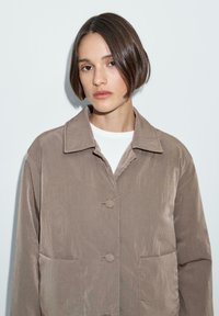 Bruine, gestructureerde jas met een kraag, twee voorzakken en knoopsluiting. Draagt over een wit shirt met een gladde afwerking.