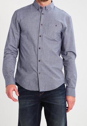 Homme portant une chemise grise à manches longues boutonnée avec une poche poitrine et un jean bleu foncé, debout devant un fond uni.