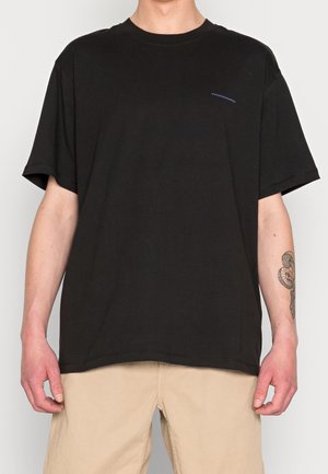 Mężczyzna w czarnej koszulce z krótkim rękawem z małym logo na lewej piersi i beżowych spodniach, z odsłoniętymi przedramionami i widocznym tatuażem na prawej ręce.