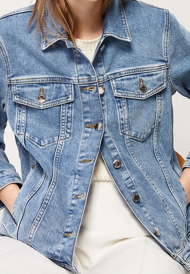 Nainen, jolla on päällään vaaleansinisestä denimmistä valmistettu takki, jossa on metalliset napit, valkoisen neulepaidan ja valkoisten housujen päällä, kädet takin taskuissa.