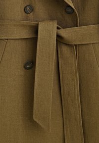 Manteau en lin marron avec ceinture en tissu assortie nouée en nœud et trois boutons foncés visibles à l’avant.