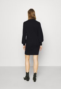 Robe en tricot noire avec des manches longues et bouffantes, silhouette ajustée et ourlet côtelé. Modèle portant des bottes noires à talons épais.