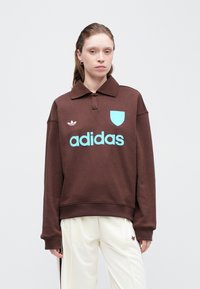 Rjav pulover s kapuco, ki ima ovratnik, z lahkotno modrim logotipom "adidas" in akcentnim ščitom. Mehka tkanina, sproščen kroj, rebrasti manšeti.