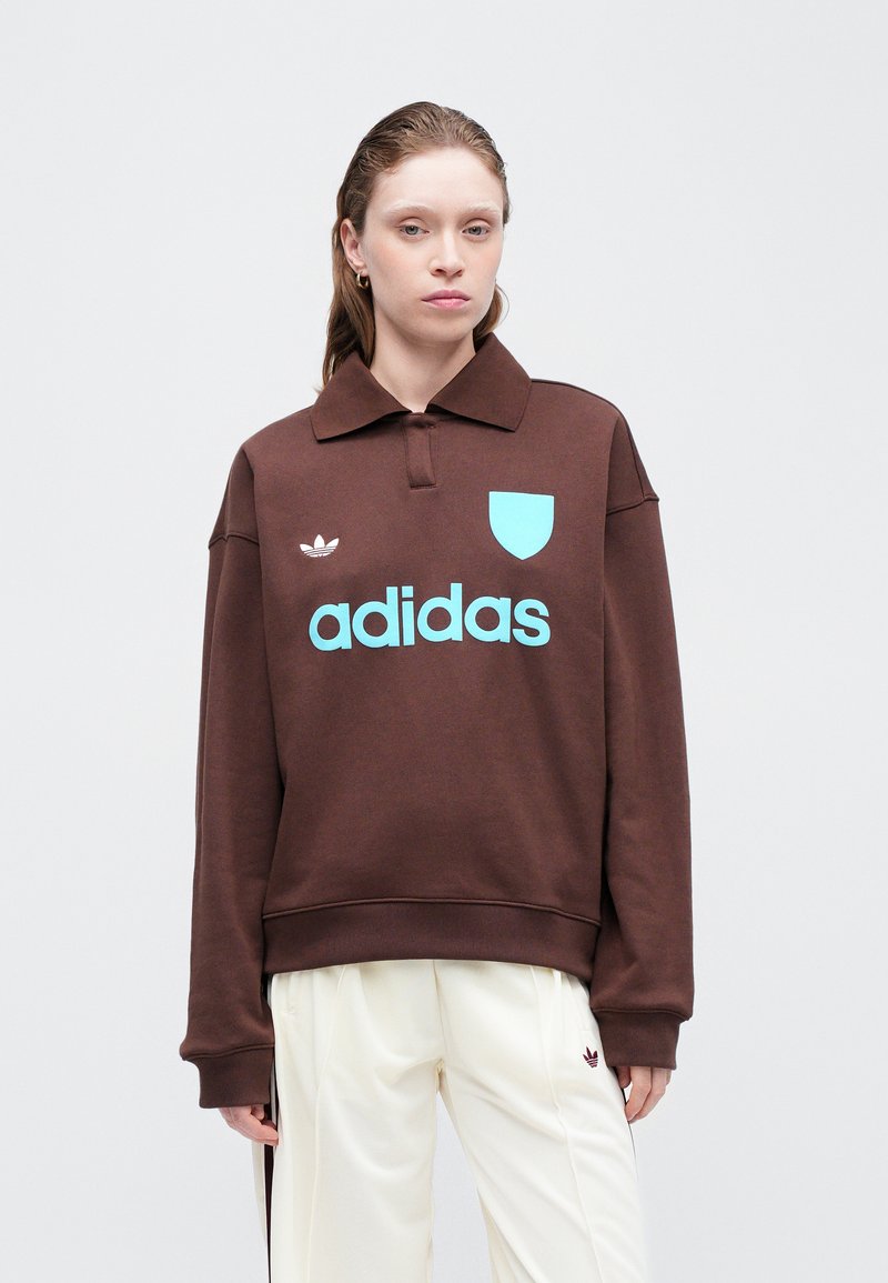 Sweatshirt marron à col, arborant un logo "adidas" bleu clair et un blason d'accent. Tissu doux, coupe décontractée, poignets côtelés.
