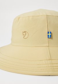 Beige bøttehatt laget av myk stoff, med et sydd revlogo og et lite svensk flagg som aksent på siden. Glatt overflate.