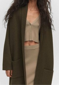 Frau mit olivgrünem Oversized-Mantel über einem beigen, kurz geschnittenen, gerippten Oberteil und passendem hochgeschnittenen Rock, gezeigt von den Schultern bis zur Hüfte.