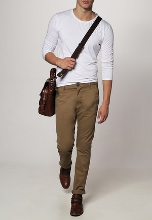 Hombre caminando con camisa blanca de manga larga, pantalones caqui, zapatos de cuero marrón y llevando un bolso de cuero marrón al hombro.