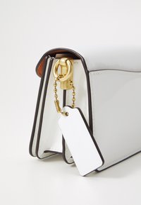 Witte lakleren tas met zwarte rand, met een gouden metalen sluiting en een decoratieve ketting bevestigd aan een lege witte label.