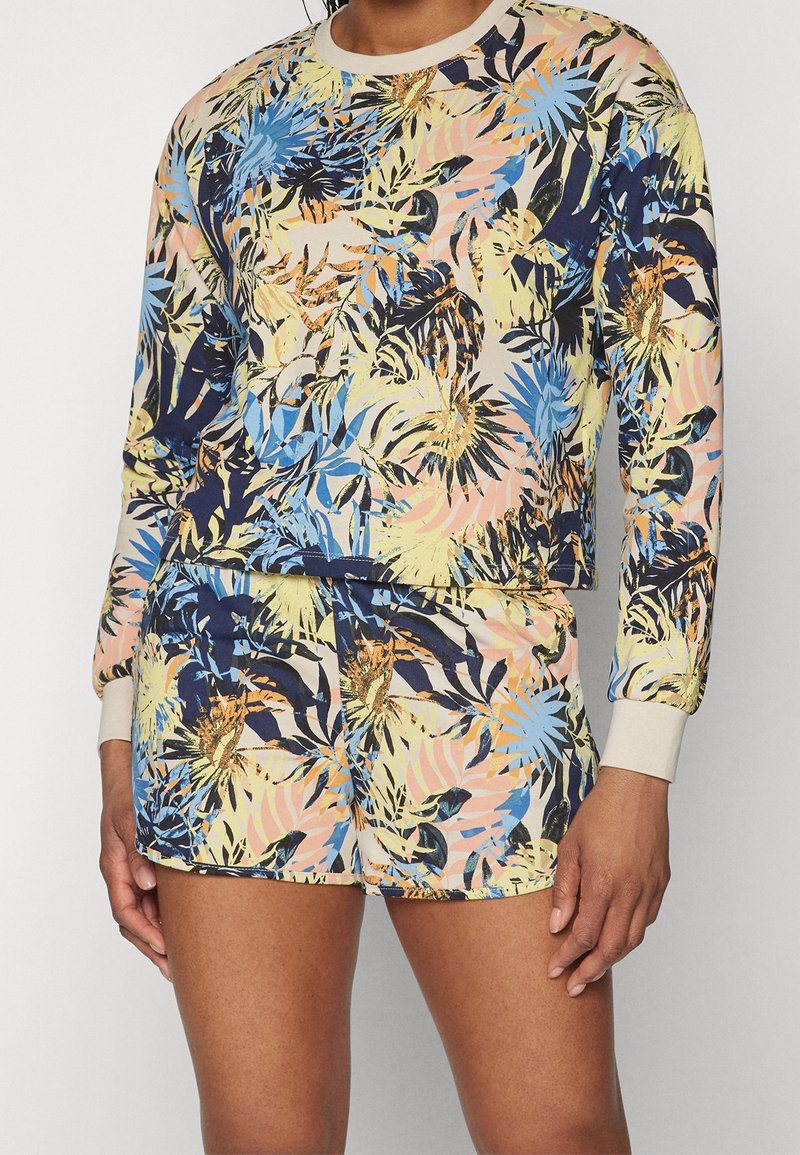 Ensemble haut à manches longues et short avec un imprimé coloré de feuilles tropicales et de fleurs en tons bleu, jaune, pêche et marine sur un fond clair.