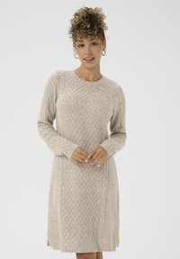Vrouw met krullend haar, draagend van oorringen en een beige gebreide lange mouwen jurk, staande tegen een effen lichte achtergrond.