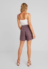 Shorts à imprimé floral avec une taille smockée en rouge foncé, ornés de motifs roses et blancs. Associés à un caraco blanc et des sandales à talons carrés beiges.