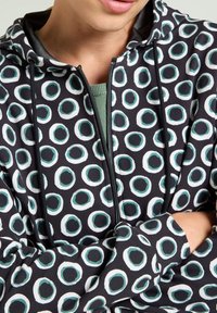 Schwarzer Kapuzenpullover mit einem sich wiederholenden kreisförmigen Muster in Weiß und Teal, mit Reißverschluss vorne, Kordelzügen und einer weichen Textur.