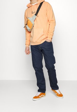 Felpa arancione con tasca frontale, abbinata a jeans blu scuro; indossa scarpe da ginnastica arancioni e bianche. Borsa a tracolla grigia e arancione indossata.