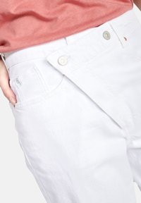 Witte katoenen broek met een unieke asymmetrische tailleband en zilveren hardware. Glad textuur met een lichte glans, zijzakken zichtbaar.