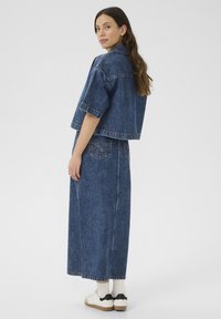 Farkkutakki, jossa on lyhyt malli ja lyhyet hihat, yhdistettynä pitkään farkkuhameeseen. Molemmat vaatekappaleet keskikokoisessa pesussa ja niissä on ompeluominaisuudet.