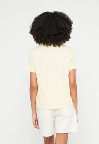 Rövid ujjú, világos sárga póló egyenes szabással és puha textúrával, fehér rövidnadrággal párosítva, hátulnézetből.