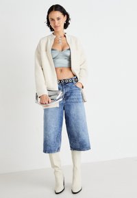Krémový, nadýchaný kardigan přes modrý saténový crop top, spárovaný s vysokými denimovými kalhotami do culottes a bílými vysokými kozačkami, držící stříbrnou kabelku.