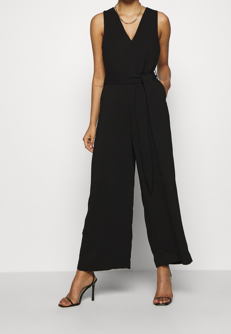 Černý bezramínkový overal s hlubokým V výstřihem, páskem v pase, širokými nohavicemi a lehkým textilním materiálem, doplněný sandály na vysokém podpatku s řemínky.