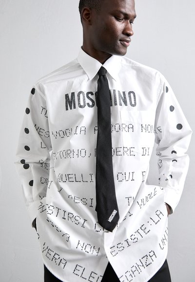 Cămașă albă cu guler, având text negru și puncte polka, cu o cravată neagră și o etichetă "Moschino" pe cravată, evidențiind un design unic.
