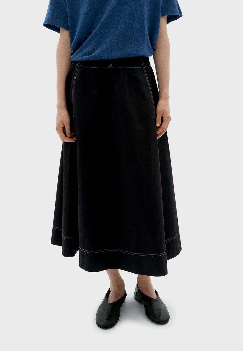 Zwarte A-lijn rok van denim, met contrasterende witte stiknaden en een tailleband met een knoop, gecombineerd met zwarte instapschoenen.