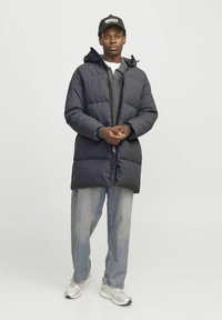 Marineblå puffjakke med hætte, lynlås og synlige sømdetaljer, parret med en grå sweater og lyse denimjeans.