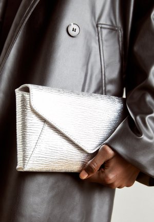 Mano de piel oscura sosteniendo un bolso de mano plateado con textura, llevado con un abrigo de cuero gris oscuro que tiene un gran botón y bolsillo.