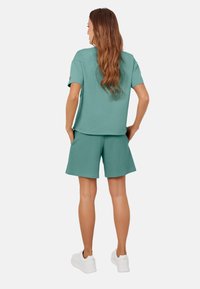 Zielona bluzka z krótkim rękawem w połączeniu z dopasowanymi szortami o fakturowej strukturze, wykonanymi z miękkiego materiału. Modelka nosi białe sneakersy, prezentując swobodny strój.