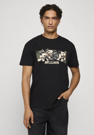 Jeune homme aux cheveux bouclés portant un T-shirt noir avec texte camo "UNFAIR ATHLETICS" et un jean noir, debout avec une main dans la poche.