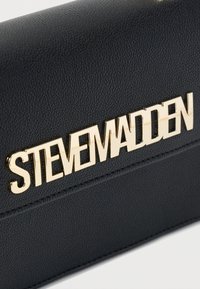 Μαύρη υφή από δέρμα με γυαλιστερά χρυσά μεταλλικά γράμματα που σχηματίζουν την ένδειξη STEVE MADDEN τοποθετημένα οριζόντια.