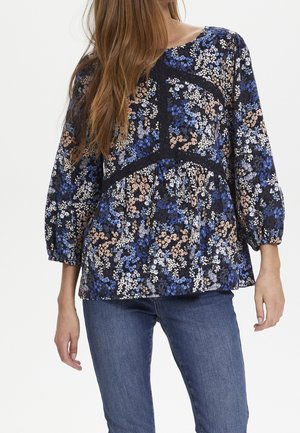 Blouse florale marine avec des motifs bleus, blancs et pêches. Présente un col rond et des manches ¾. Texturée avec une petite bordure en dentelle.