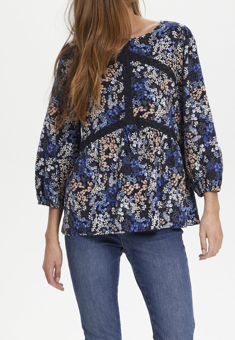 Blouse florale marine avec des motifs bleus, blancs et pêches. Présente un col rond et des manches ¾. Texturée avec une petite bordure en dentelle.