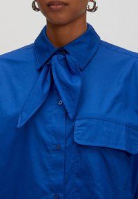 Blauw overhemdd met een strik om de hals, voorzien van een knopenlijst en een borstzak. Gladde, geweven stof met een glanzende afwerking.