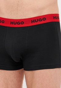 Fekete boxer alsónadrág piros elasztikus derékpánttal, amelyen a "HUGO" felirat fekete színben található. Az anyag simának és testhezállónak tűnik.