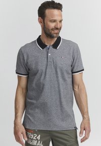 Siva polo majica z črnim ovratnikom in belimi obrobami. Ima dva gumbka pri vratu in majhen barvno razdeljen logotip na prsih.