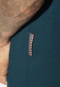 Nahaufnahme eines Arms, der neben einem dunkelblauen Stoff ruht, auf dem ein kleines "Napapijri"-Etikett mit roten und weißen Streifen zu sehen ist.