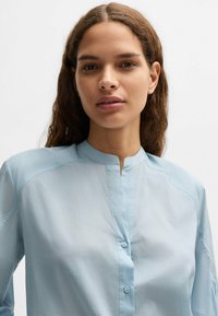 Jasnoniebieska koszula zapinana na guziki z kołnierzem band, wykonana z gładkiego materiału. Posiada luźny krój i subtelne przeszycia na ramionach.