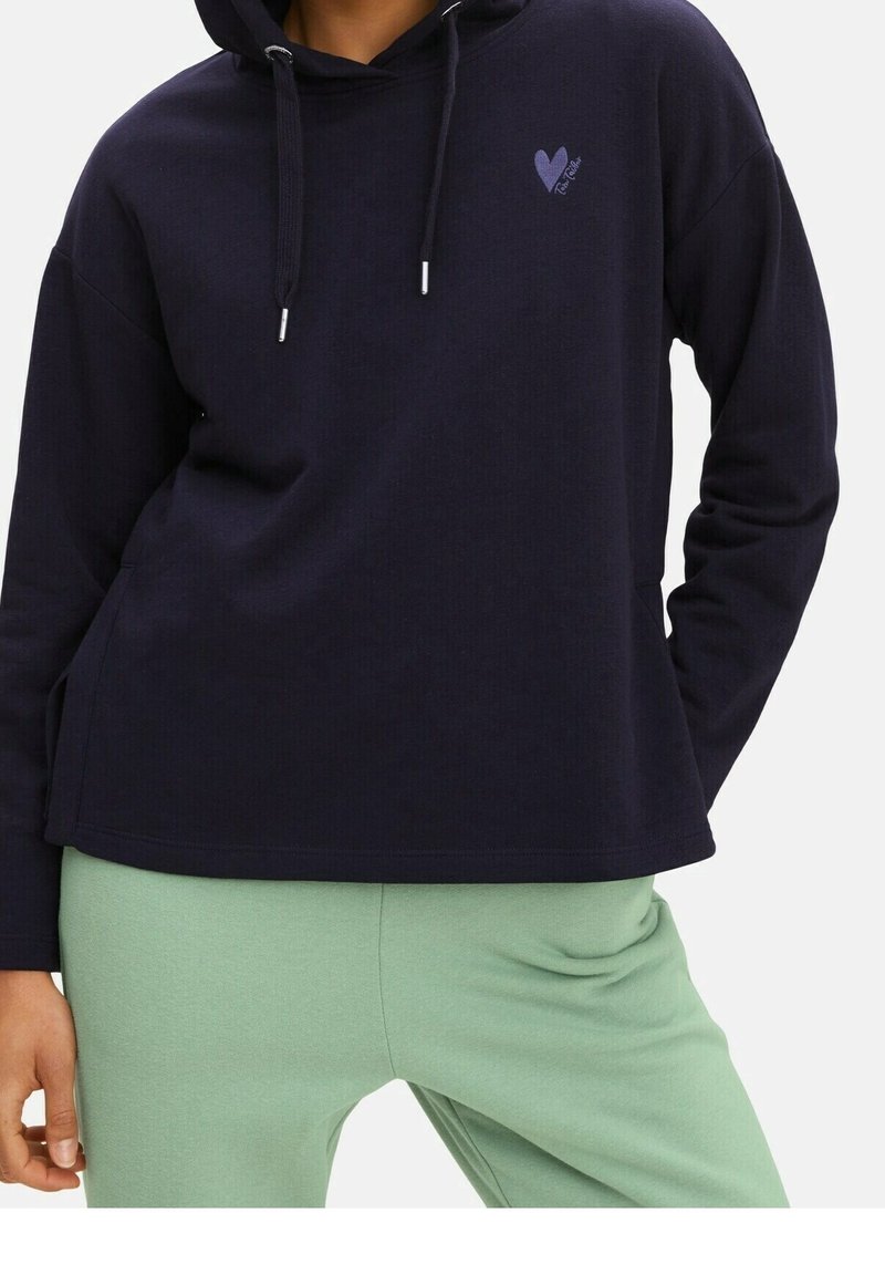 Personne portant un sweat à capuche ample bleu marine avec cordons et un petit motif cœur, associée à un pantalon vert clair.