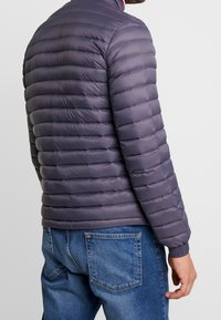 Giubbotto imbottito di un viola scuro con cuciture orizzontali. Presenta un collo alto e polsini a coste, abbinato a jeans blu. Finitura opaca.