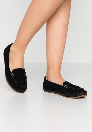Persoon draagt zwarte suède mocassin loafers met kwastjes en stikseldetails, staand op een lichtgekleurde vloer.