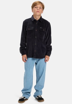 Marineblauwe corduroy shirt met knoopsluiting aan de voorkant, twee borstzakken en een casual pasvorm, gecombineerd met lichtblauwe jeans en zwarte sneakers.