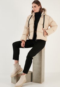Beige puffer jacket with a hood, zipper front, and black drawstrings. Paired with black pants and beige ankle boots with a chunky sole.