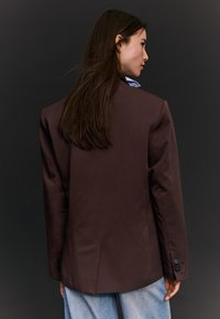 Ženska z dolgimi temnimi lasmi, oblečena v rjav blazer in svetlo modre jeans hlače, obrnjena bočno na temnem ozadju.