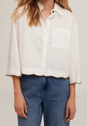 Femme portant une chemise blanche à boutons avec des bordures perlées aux manches et à l'ourlet, associée à un jean en denim bleu.