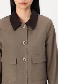Brun kragejakke med fire knapper, to brystlommer og en glat tekstur. Fargen er nøytral beige, med en kontrasterende mørk krage.