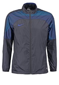 Chaqueta cortavientos de Nike en gris oscuro con rayas horizontales azules en los hombros. Presenta un cuello alto y cremallera frontal completa.