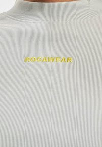 Lys grå rutet høyhalset topp med brodert "ROCAWEAR"-logo i gult. Nærbilde som fremhever tekstur og design.
