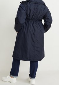 Marineblauwe lange pufferjas met een trekkoord in de taille, afneembare capuchon en ritszakken, gecombineerd met opgerolde donkere spijkerbroeken en witte sneakers.