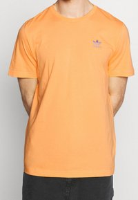 Krátký rukáv oranžové bavlněné tričko s kulatým výstřihem, na levé straně hrudi s fialovou vyšitou logem. Hladká textura.