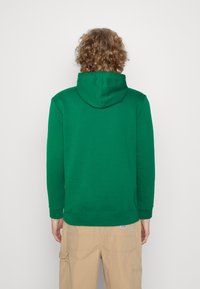 Sudadera verde con capucha, mangas largas y puños acanalados. Hecha de tela suave, diseño minimalista, vista trasera lisa sin logotipos visibles.