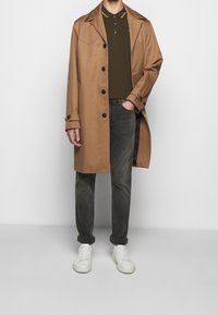 Mężczyzna w kamelowym płaszczu, ciemnozielonym polo, szarych, podwiniętych dżinsach i białych trampkach, stojący na tle jasnego, jednolitego tła.